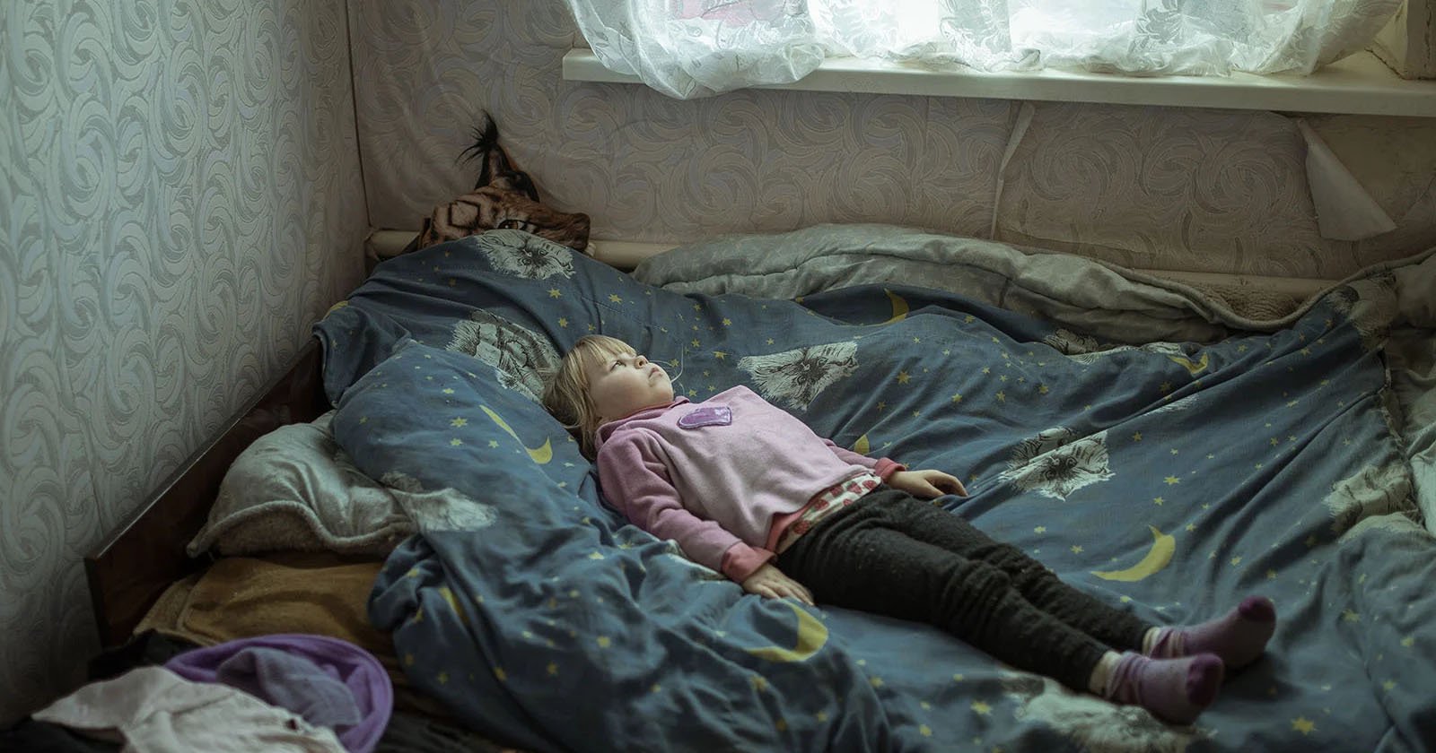 A child wearing a pink sweater and dark pants lies on a bed with a blue, moon-and-star patterned duvet. The room has light wallpaper, a window with sheer curtains, and a small stuffed toy peeks out beside the pillow.