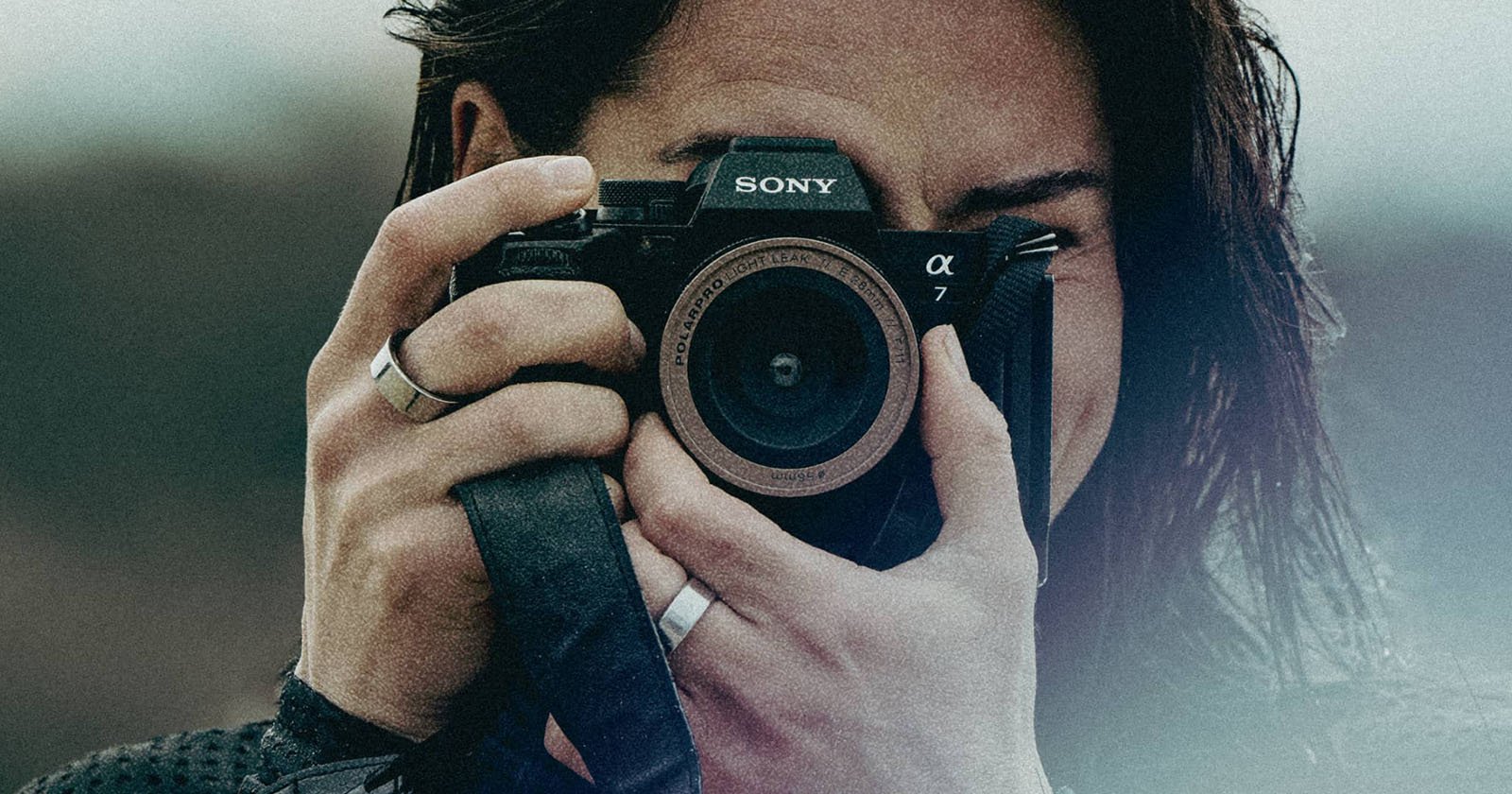 A person is holding up a Sony α7 camera close to their face, ready to take a photograph. The lens is prominently centered in the frame, and the person's fingers can be seen gripping the camera. The background is blurred, focusing attention on the camera.