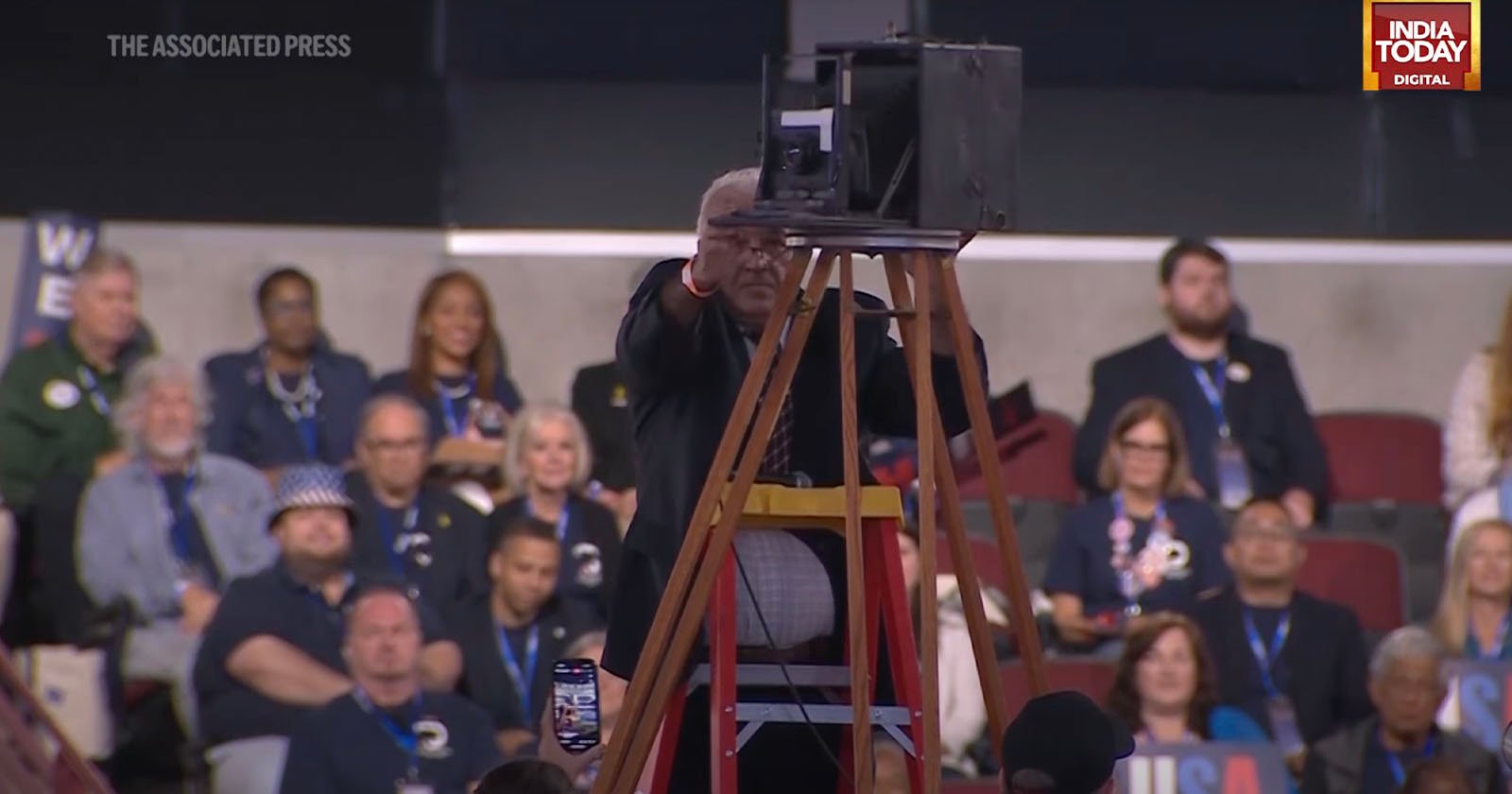 A person stands behind a large camera mounted on a tripod, addressing an audience at an indoor event. The audience is seated, with some individuals wearing badges and lanyards. A logo of India Today Digital is visible in the top right corner.