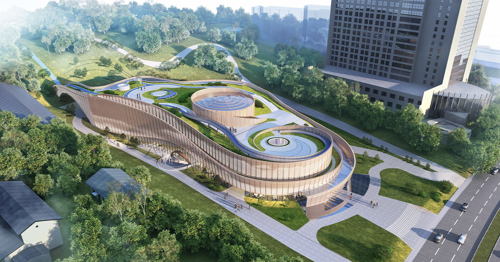 Aerial view of a modern architectural structure with flowing curves and green roofs, set in a lush park area. Surrounding roads, trees, and a tall building are visible. Pathways and landscaped areas wind around the building, integrating it into the natural environment.