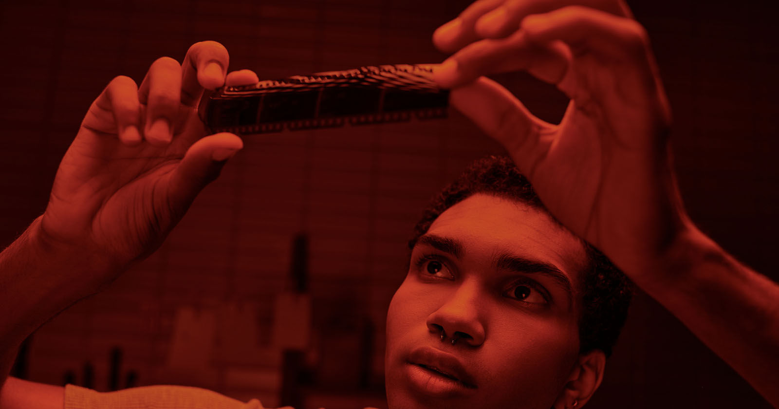 A person is examining a strip of photographic film held up to the light in a dim, red-lit room, likely a darkroom. Their expression appears focused and thoughtful as they inspect the film. The setting suggests a process related to photo development.