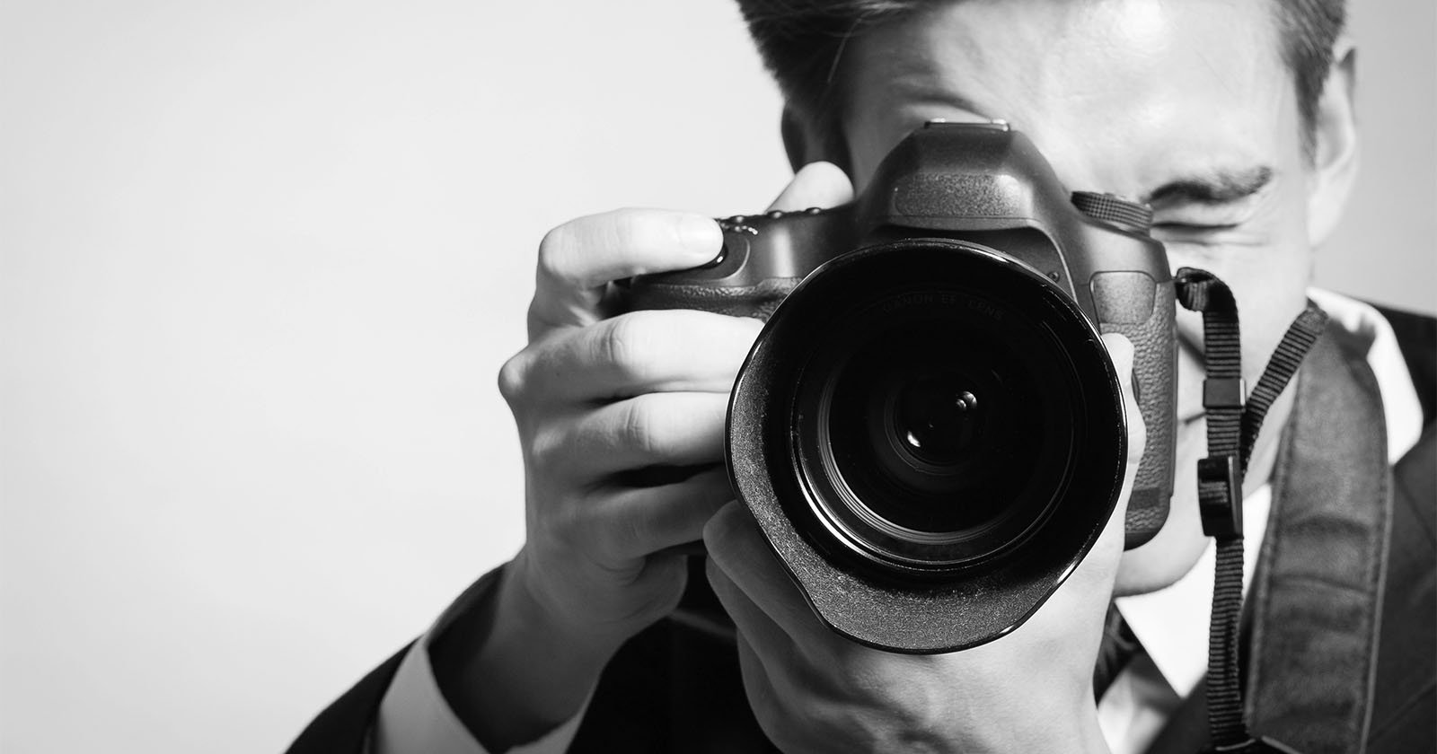 A person in a suit holding a DSLR camera up to their face, looking through the viewfinder and preparing to take a photo. The image is in black and white.