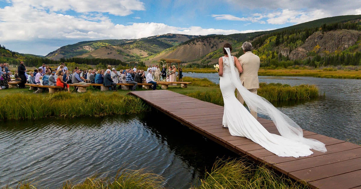 A bride in a white dress and veil walks down a wooden boardwalk next to a man in a suit towards an outdoor gathering. The setting features a picturesque lake and mountains in the background, with seated guests on both sides of the aisle.