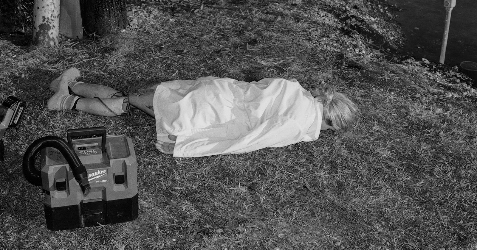 A person lies on the ground face down in a grassy area, covered with a white sheet. A vacuum cleaner is placed nearby. The scene is outdoors, surrounded by trees and foliage. The image is in black and white.