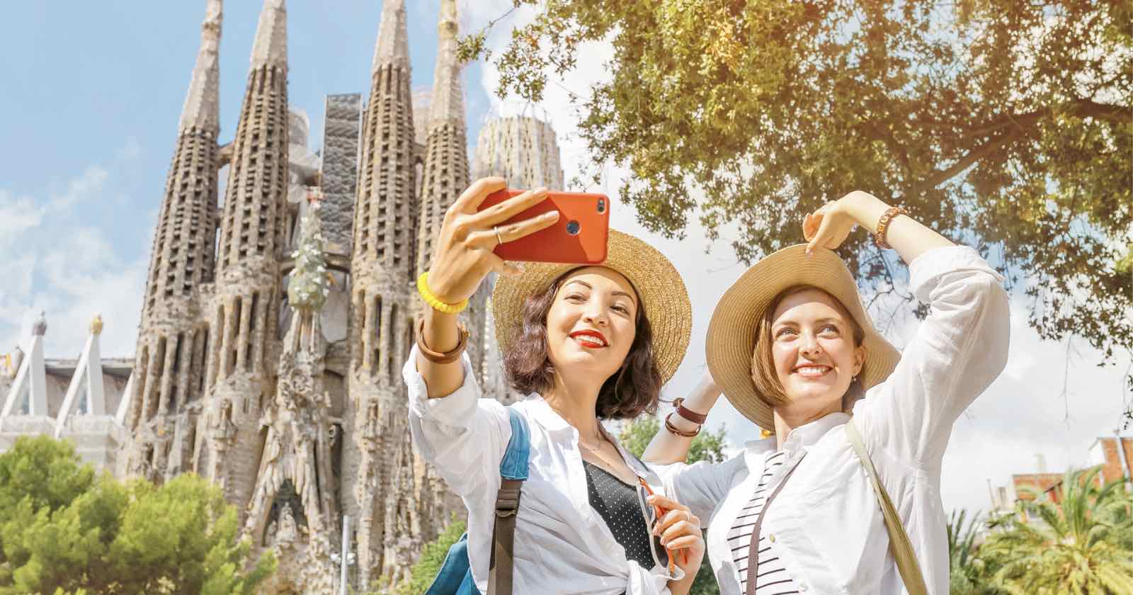 Two people in hats take a selfie with a smartphone in front of the Sagrada Família in Barcelona. They are smiling and appear to be enjoying a sunny day, with trees and the iconic church spires in the background.