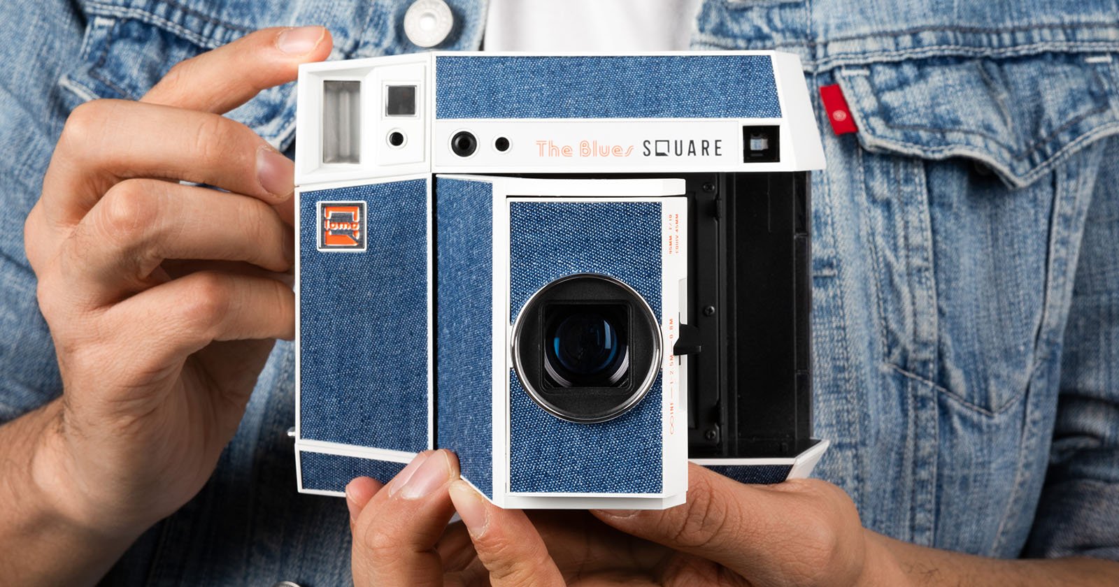 A person in a denim jacket holds a retro-style camera with a blue denim finish labeled The Blues Square. The camera has a viewfinder, lens, and stylized branding resembling a vintage instant camera.