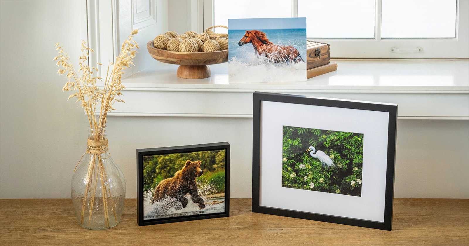 A wooden table displays framed photos of a bear in water, a white bird among green leaves, and an unframed photo of a horse running in surf. A glass vase with dried grass sits beside the photos by a window.