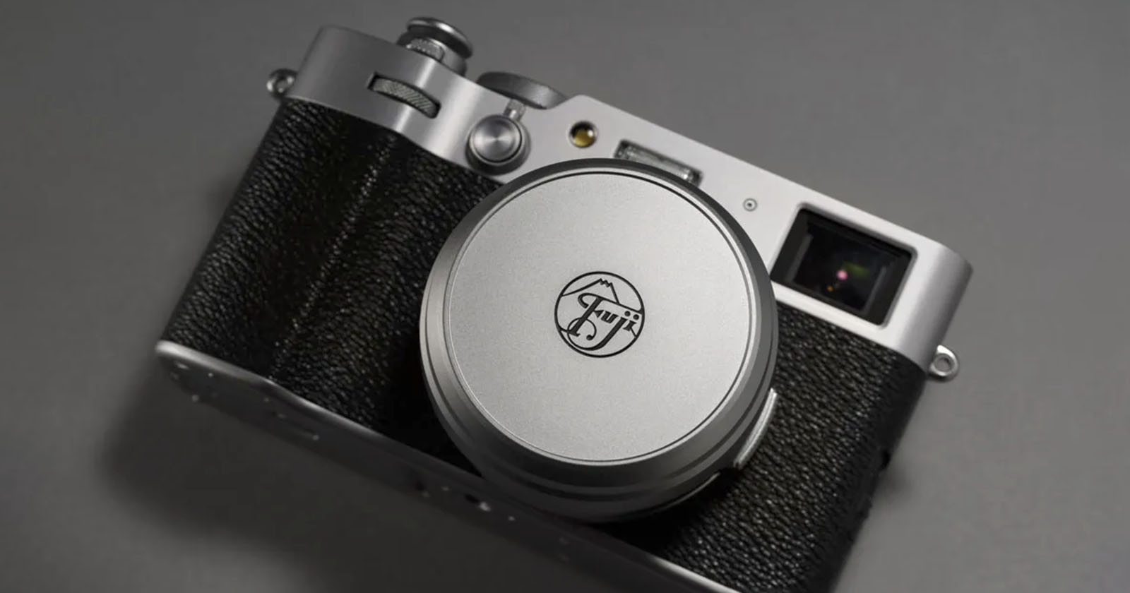 A silver and black Fujifilm camera lies on a gray surface. The camera features a retro design with a round lens cap displaying the Fujifilm logo. The body has a textured grip and various control dials visible on the top.