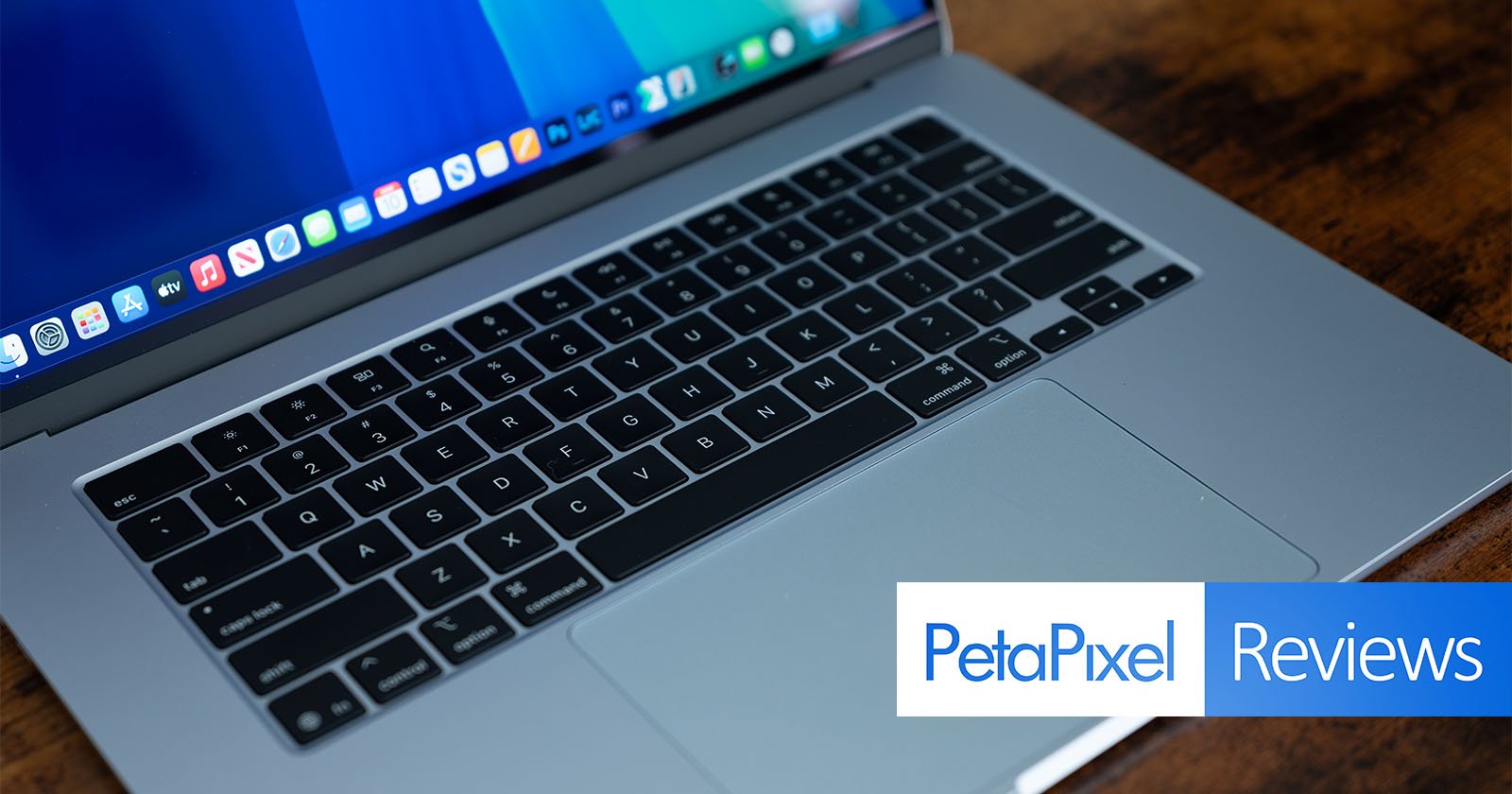 A silver laptop is open on a wooden surface, displaying a desktop screen with app icons. The keyboard and trackpad are visible. In the corner, there is an overlay with the words PetaPixel Reviews.