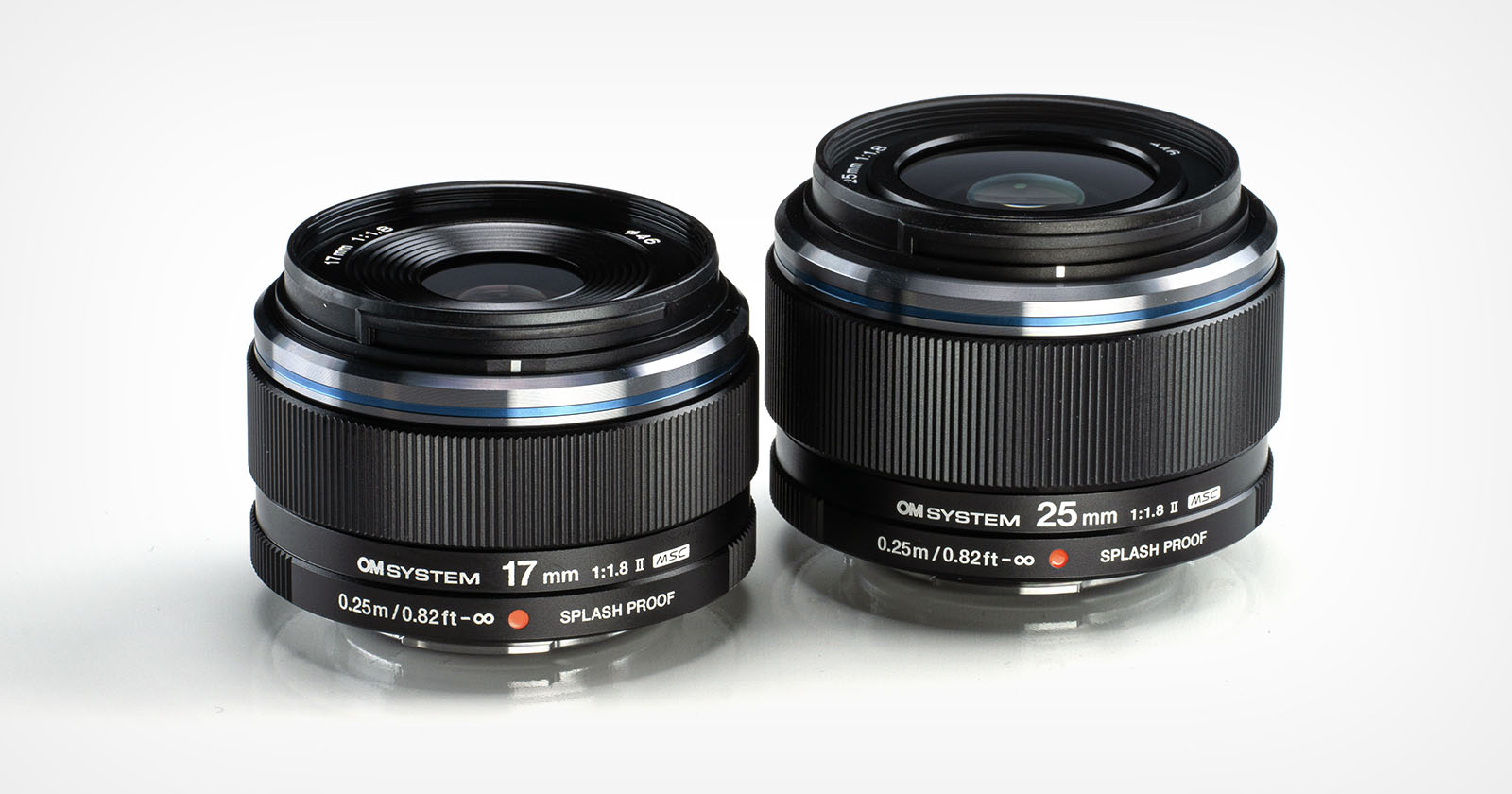 Two black camera lenses, labeled OM SYSTEM, are displayed side by side. The lens on the left is a 17mm f/1.8 and the one on the right is a 25mm f/1.8. Both lenses feature SPLASH PROOF labeling and have a sleek design.