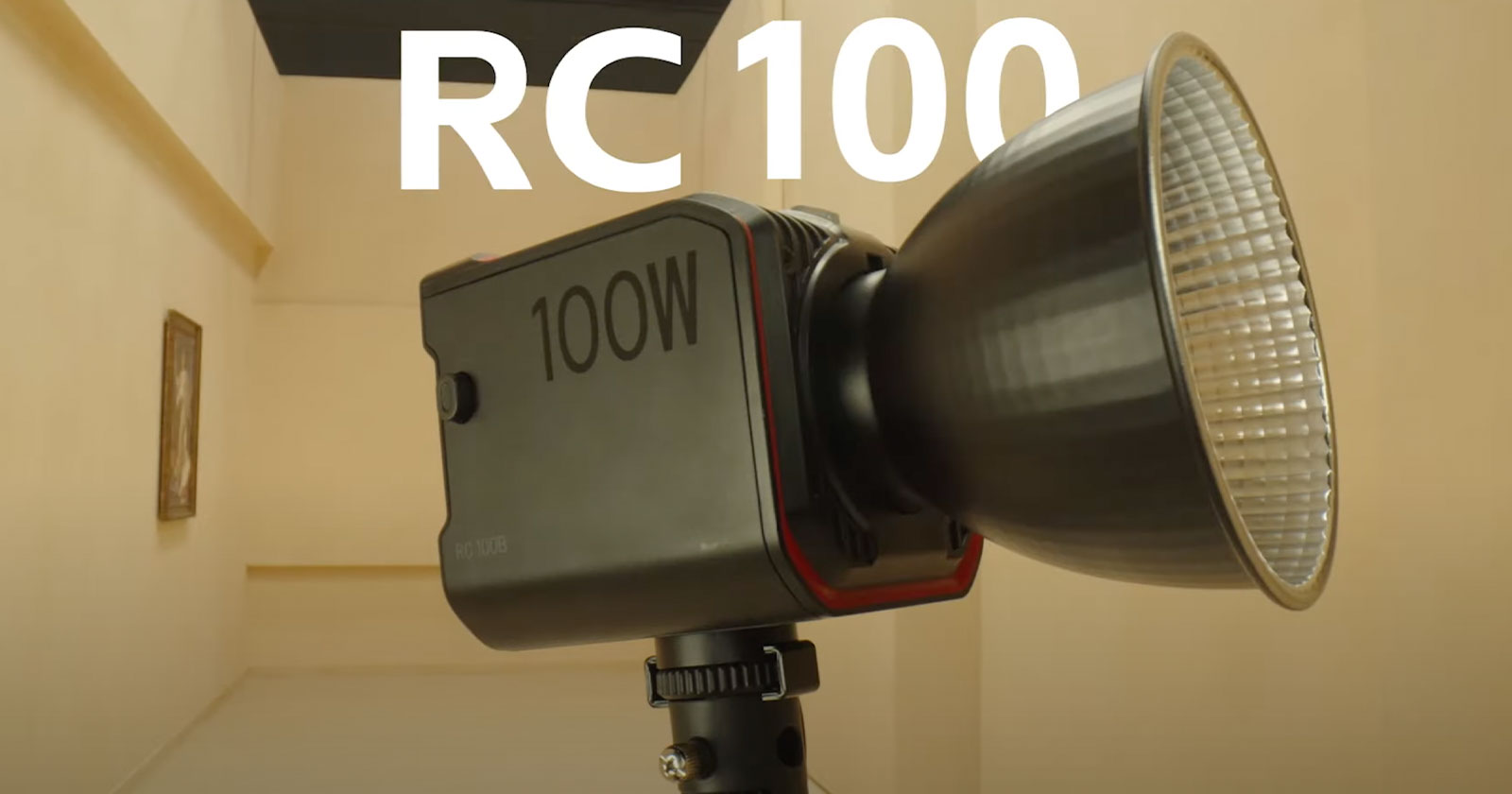A close-up image of an LED studio light labeled 100W mounted on a tripod, with the large reflective dish facing right. The background consists of beige-colored walls and a small framed picture. The bold text RC 100 is prominently displayed at the top.