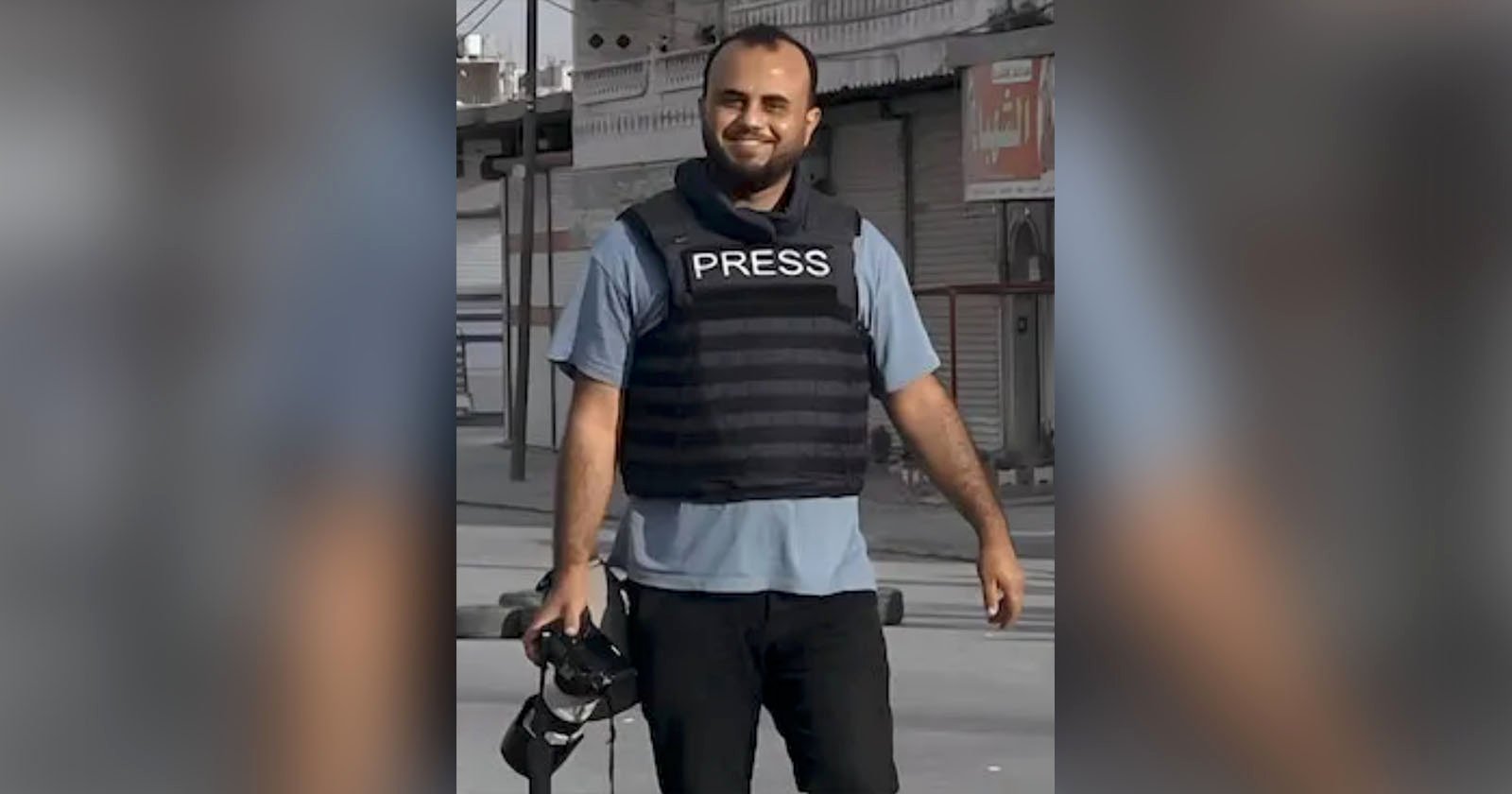 A man wearing a blue shirt and black bulletproof vest labeled PRESS stands outdoors, smiling, and holding a camera. The background features closed shop fronts and a gray street. He appears to be in a professional setting, possibly reporting or documenting an event.