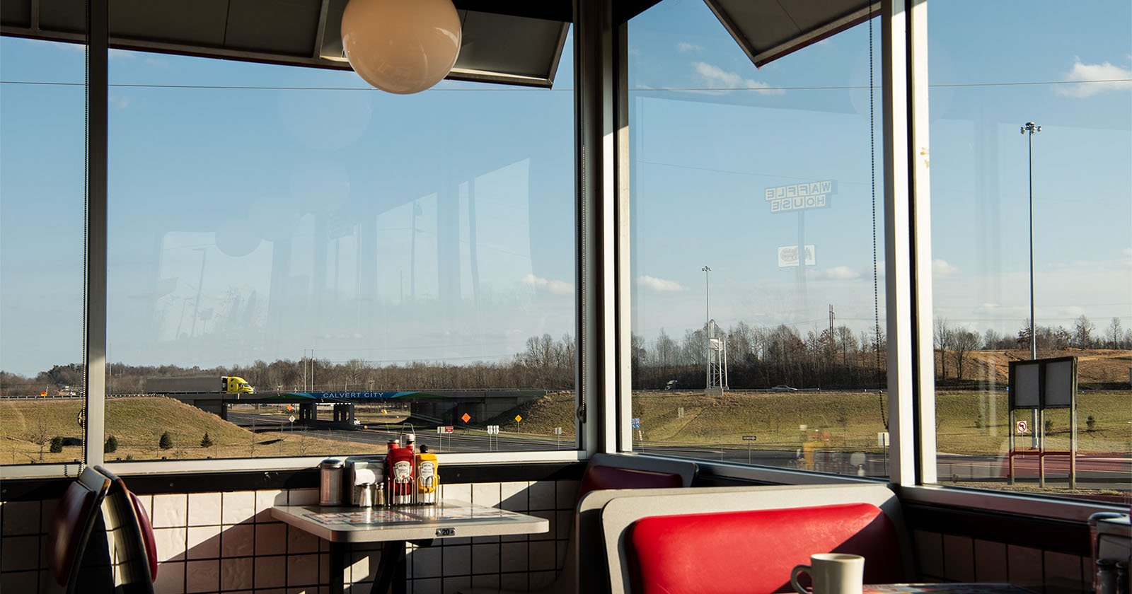 Inside a diner with red booths and a condiment-filled table by the window, overlooking a highway, grassy fields, and a yellow truck passing by under a clear sky.