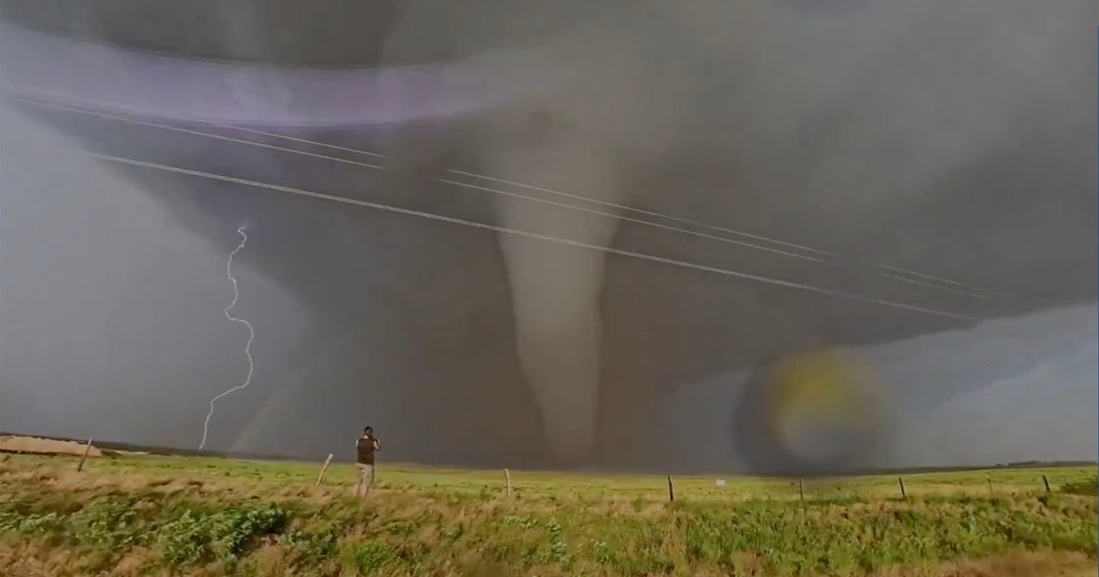 A person stands in a grassy field photographing a large tornado under a dark, cloudy sky. Lightning strikes in the background, and power lines stretch across the scene.
