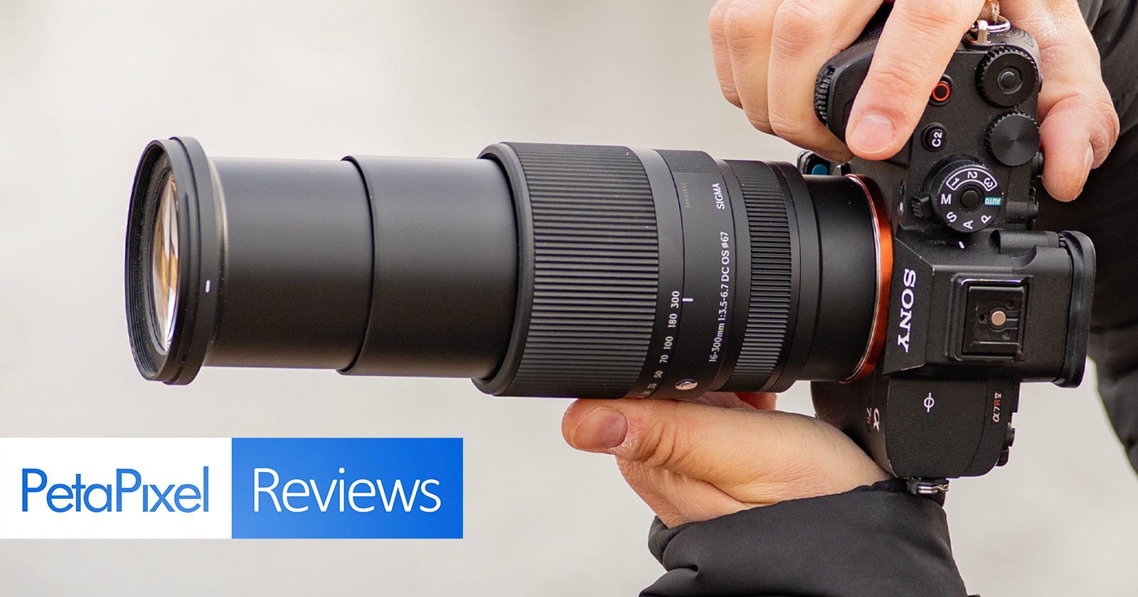 Close-up of hands holding a black Sony camera with a long telephoto lens attached. The camera is pointing to the left. Text in the bottom left corner says PetaPixel Reviews.