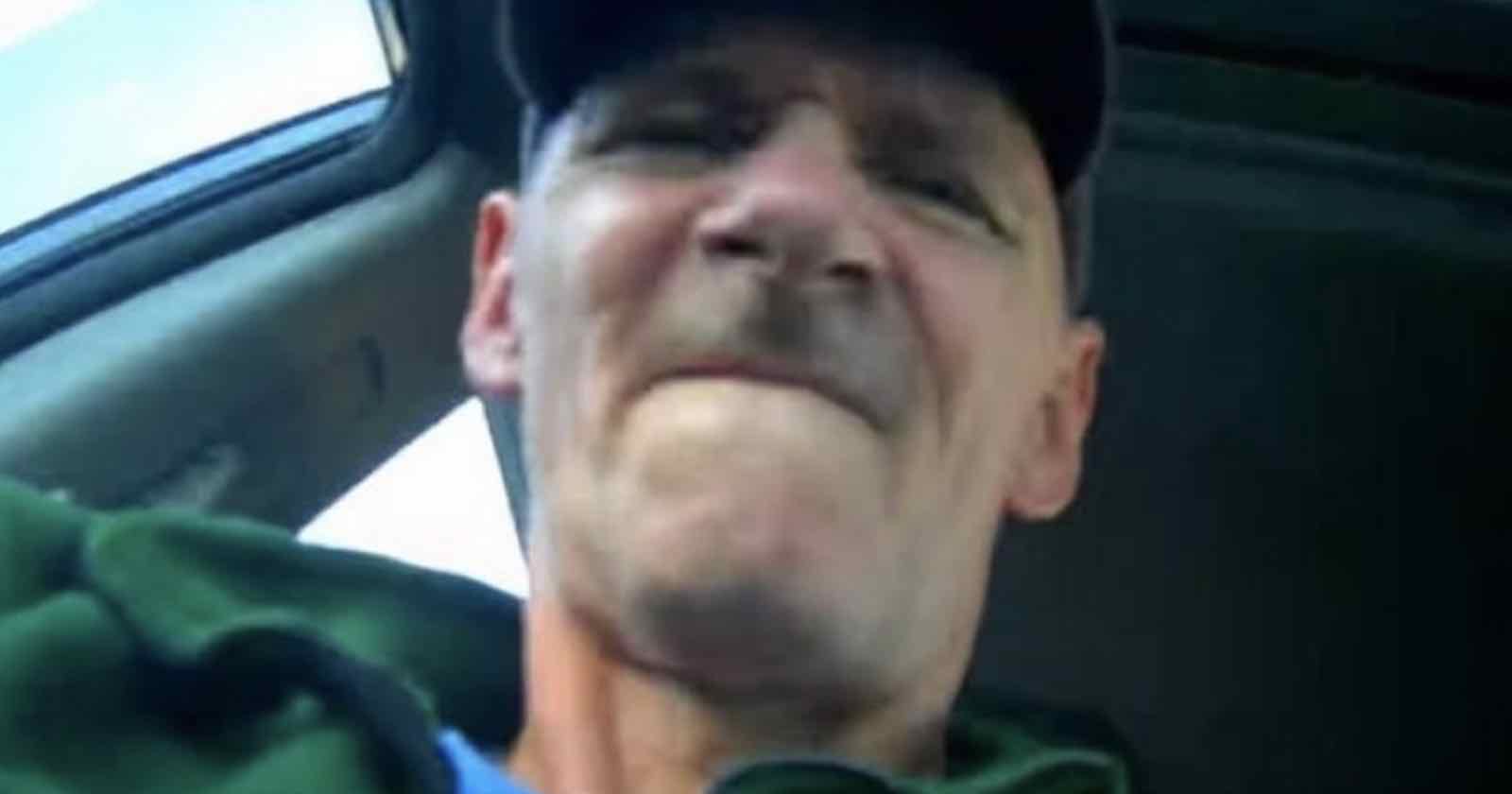 A person sitting in a car, wearing a dark cap and green jacket, with a serious expression. The image is taken from a low angle with the car window slightly open and the sky visible outside.