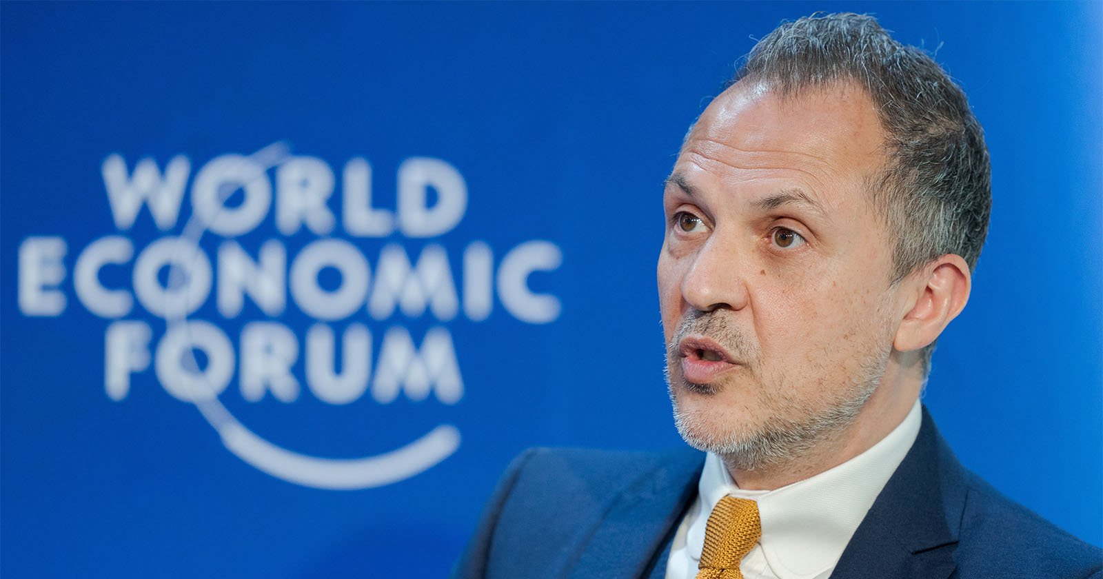 A man in a suit and yellow tie speaks in front of a blue background with the World Economic Forum logo.