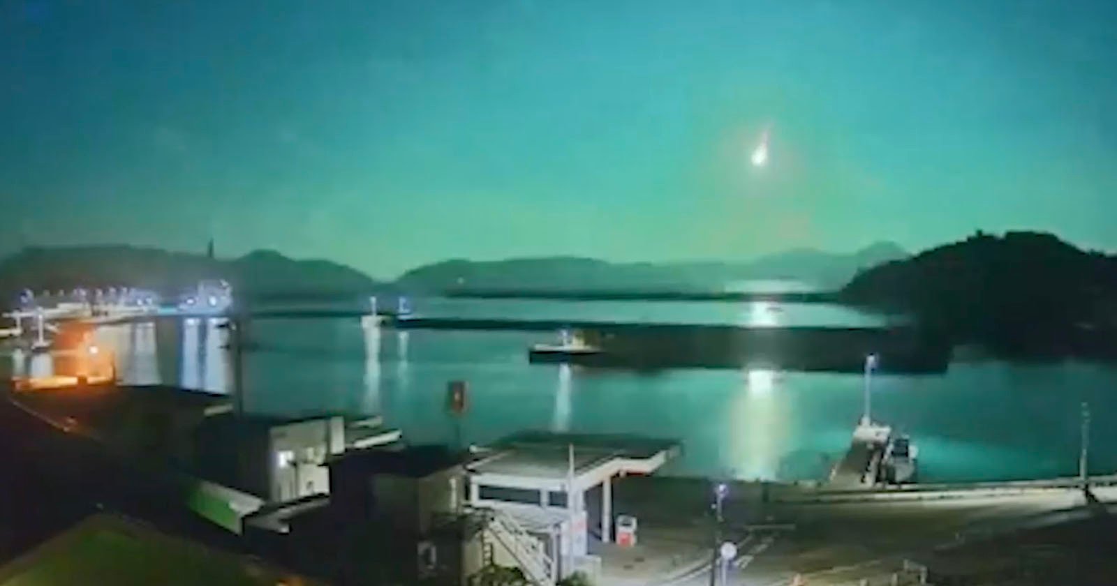 A bright meteor streaks across the night sky above a calm waterfront with buildings, docks, and distant hills illuminated by moonlight or city lights.