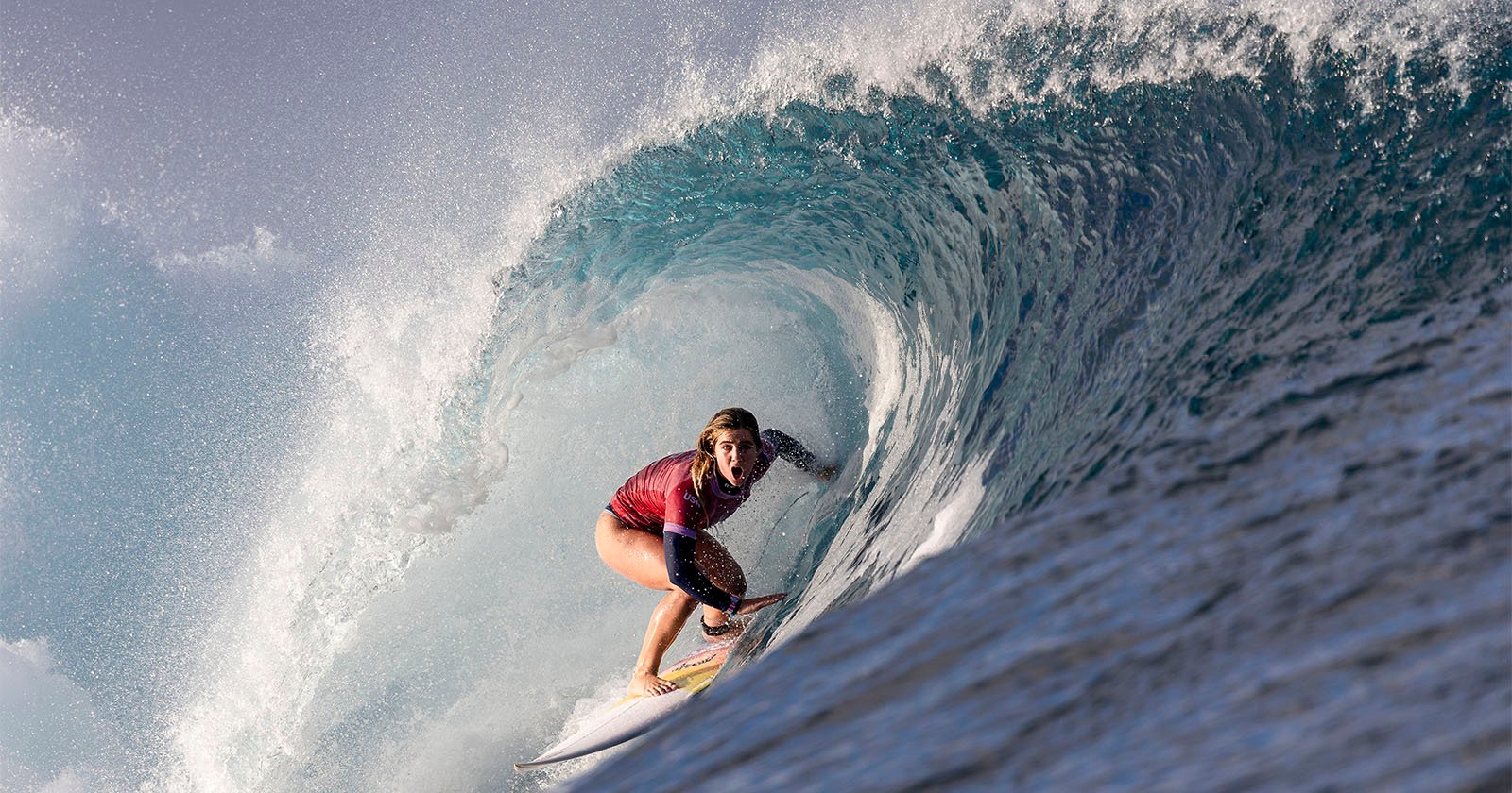 A surfer in a red shirt crouches low on their board, riding inside a massive, curling ocean wave. The water forms a tunnel around the surfer, capturing a dynamic and dramatic action moment. The sky is partly cloudy, adding to the intensity of the scene.