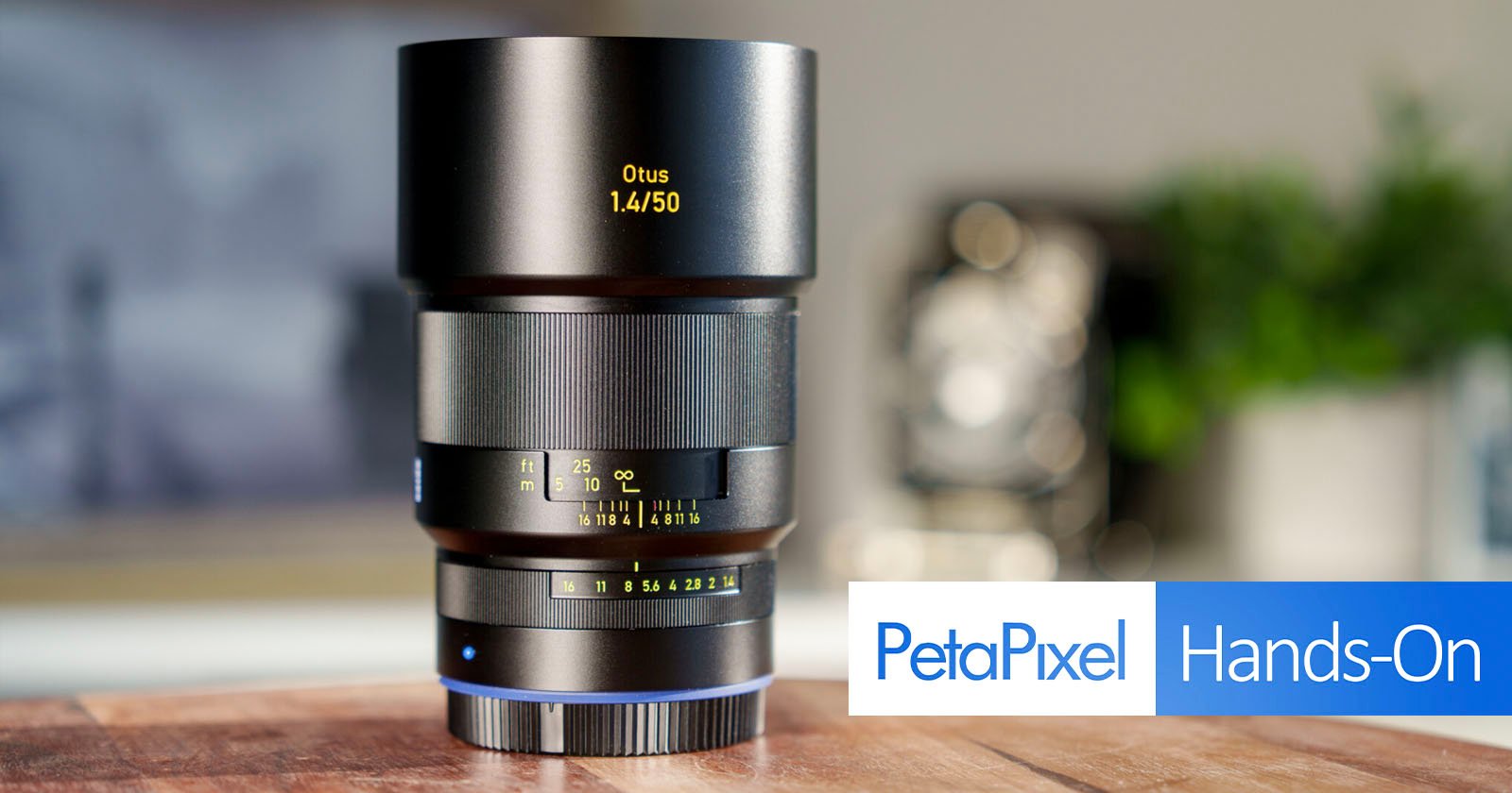 A Zeiss Otus 1.4/50 camera lens on a wooden surface. The lens is in focus with visible aperture and focus markings. The background is blurred, and there's a PetaPixel Hands-On graphic at the bottom right.