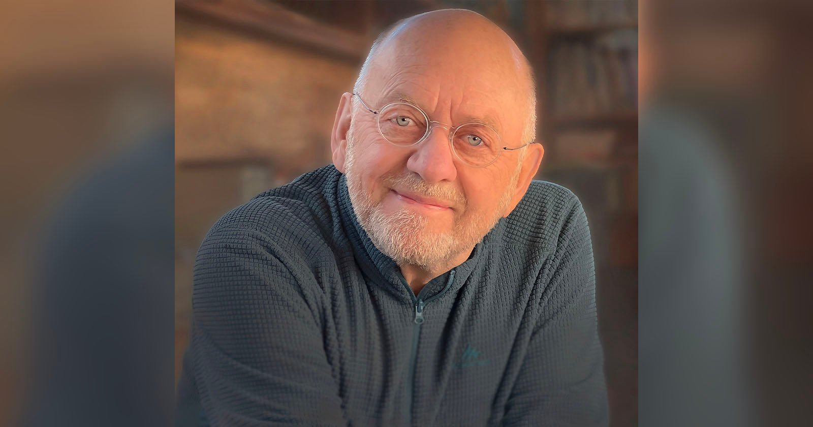 Elderly man with glasses and a beard smiles warmly. He's wearing a textured dark sweater, sitting indoors against a softly blurred background of shelves and warm lighting.