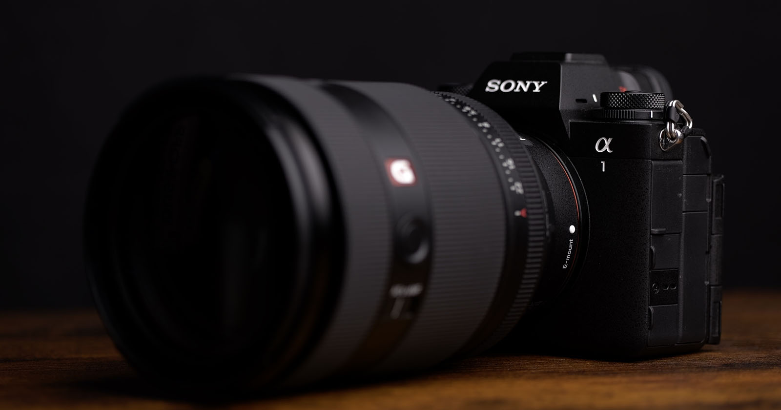 Close-up of a black Sony camera with a large lens on a dark background. The camera appears to be positioned on a wooden surface.