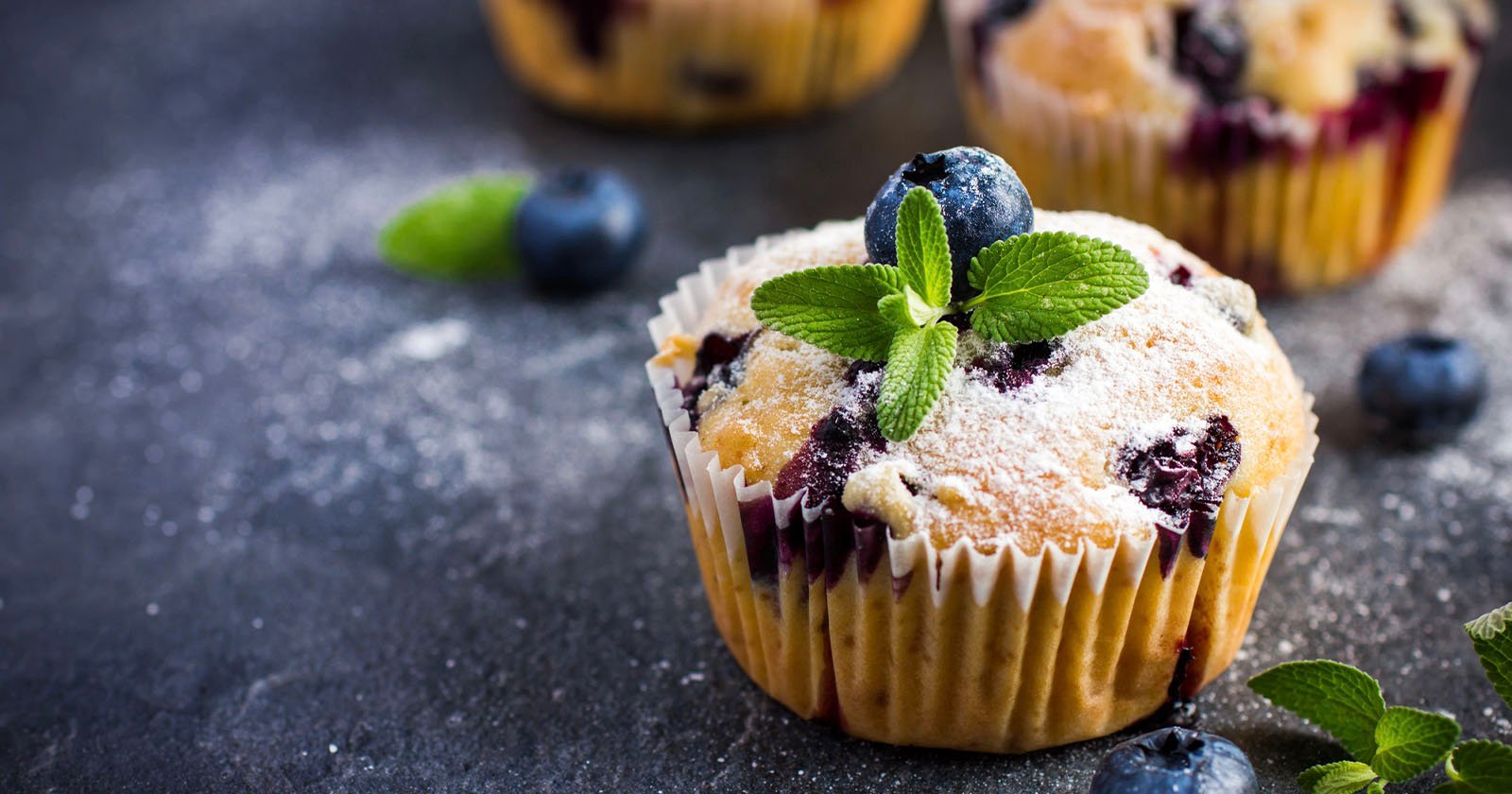 A blueberry muffin topped with powdered sugar, a fresh blueberry, and mint leaves sits on a dark surface, surrounded by scattered blueberries and mint.