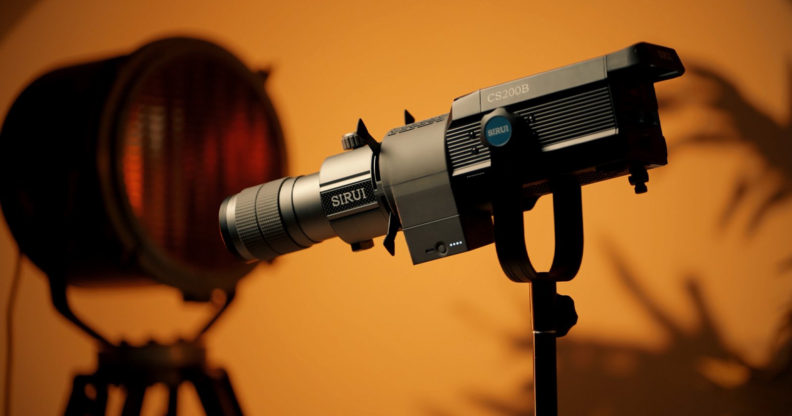 A professional camera with the brand name SIRUI is mounted on a tripod, facing left. It is set up in a studio with warm, ambient lighting, and a blurred background featuring a large spotlight. Shadows of plants can be seen on the wall.