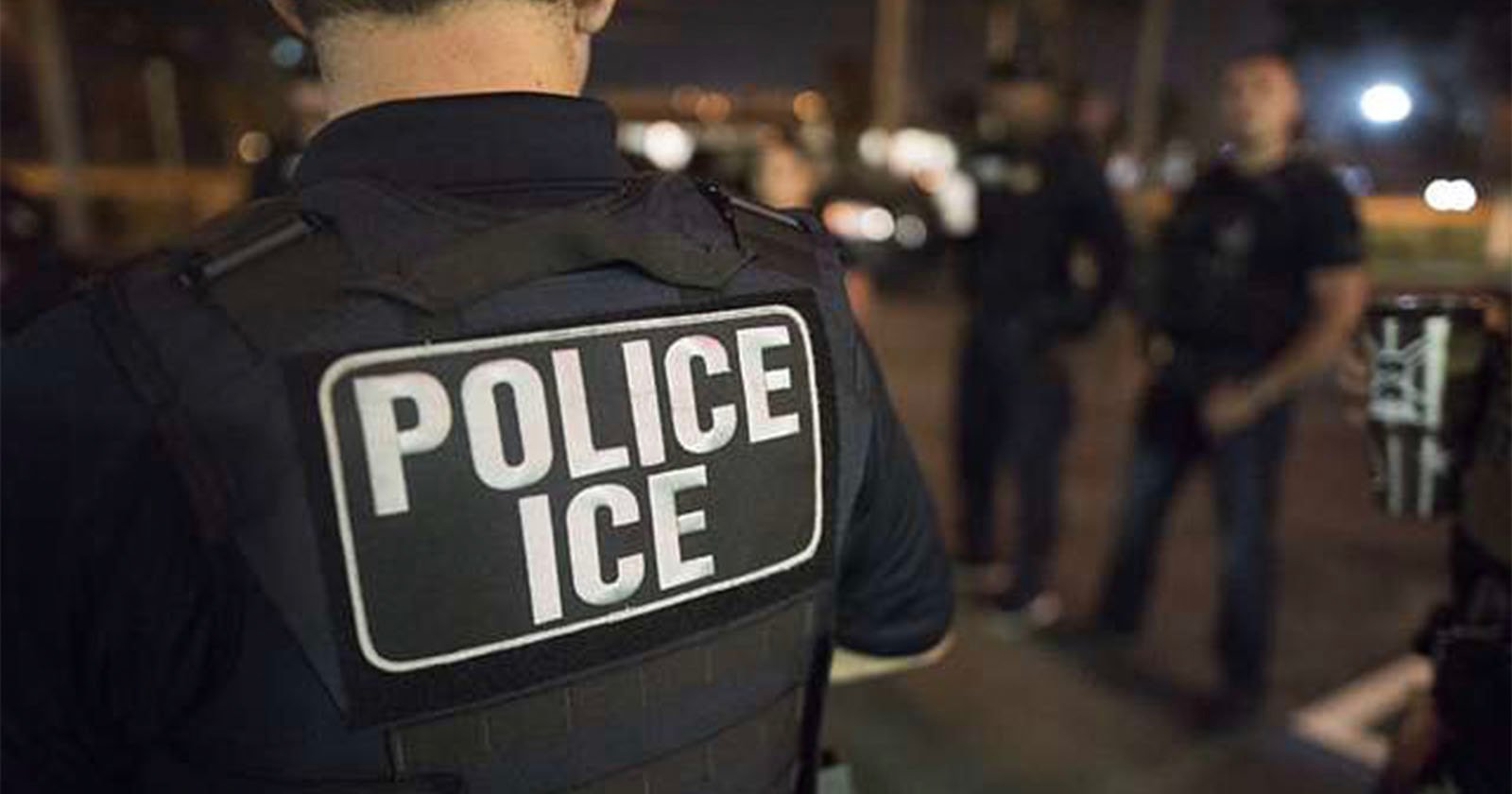 A person wearing a vest labeled Police ICE stands in the foreground at night, with several other people blurred in the background under streetlights.
