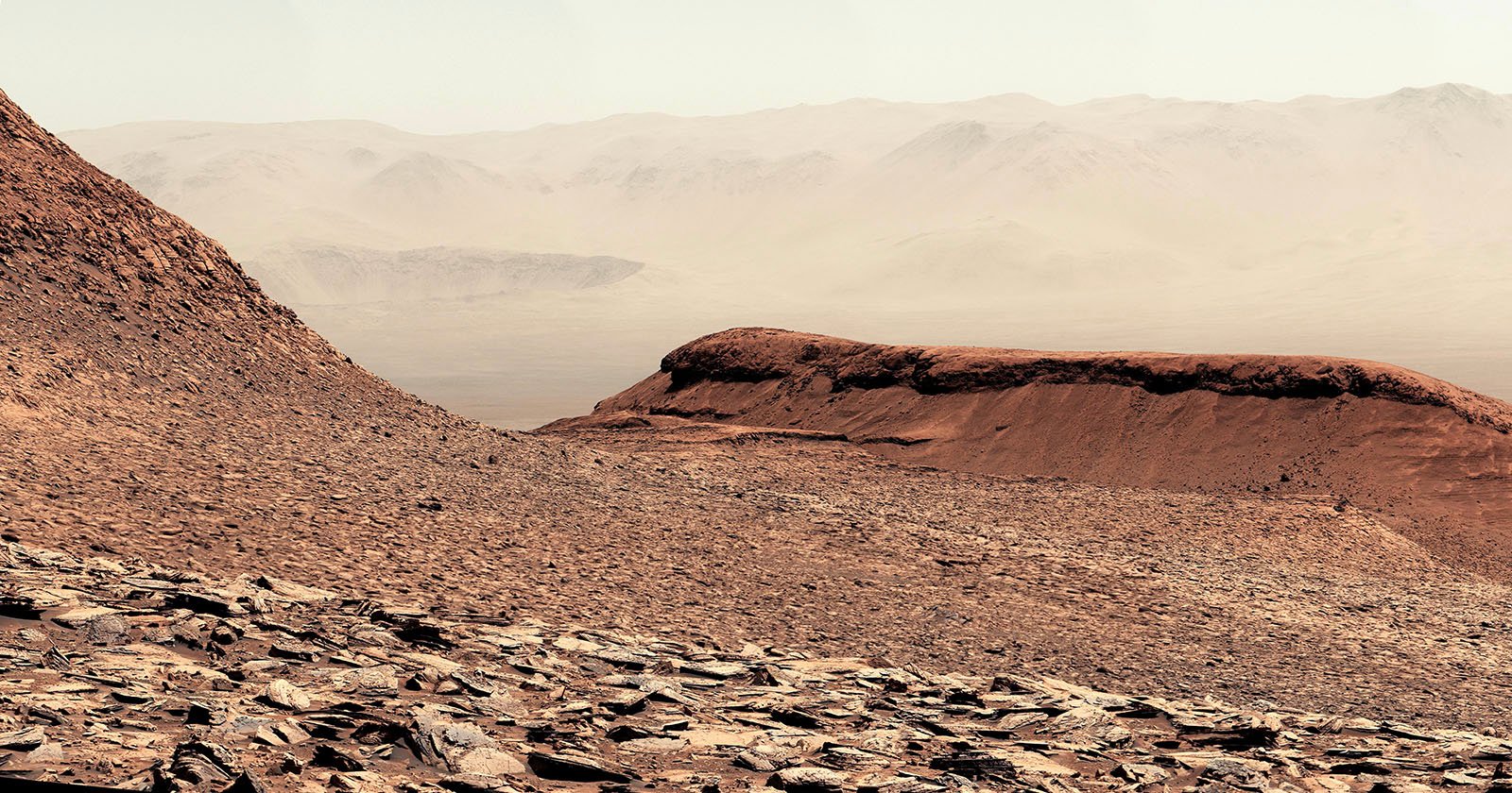 A rocky, reddish-brown Martian landscape with layered hills and mountains fades into a hazy, distant horizon under a pale sky. The terrain is dry and rugged, covered in loose rocks and dust.