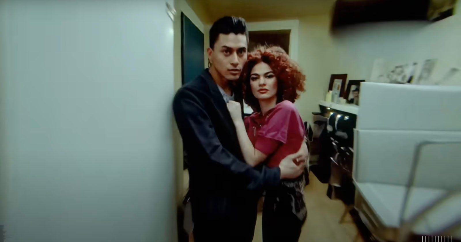 A man and a woman embrace in a narrow hallway, displaying intense expressions. the man wears a dark blazer, and the woman has voluminous curly red hair.