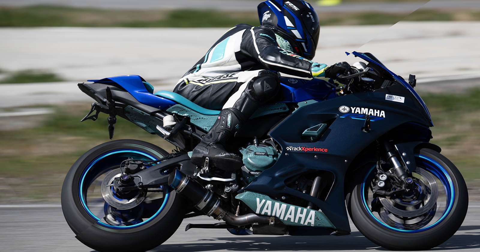 A motorcyclist in full racing gear and helmet is riding a blue Yamaha sports bike on a racetrack. The rider is leaning into a turn, demonstrating an aggressive racing posture. The background is slightly blurred, indicating high speed.