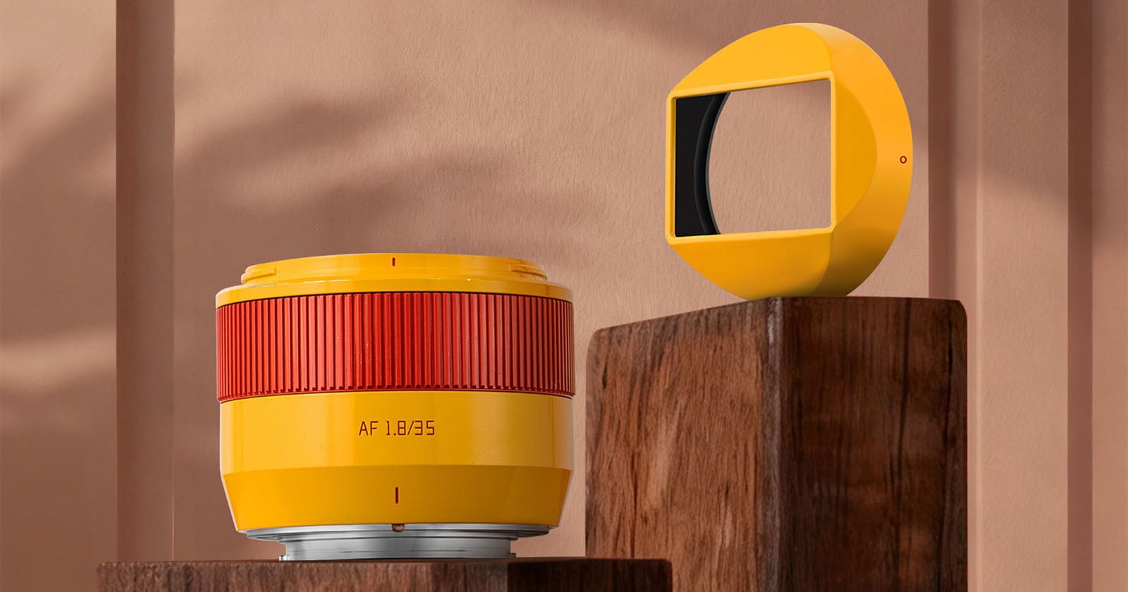 A yellow camera lens with a red ring and AF 1.8/85 text sits on a wooden block next to a matching yellow geometric object with a rectangular black opening, against a beige background.