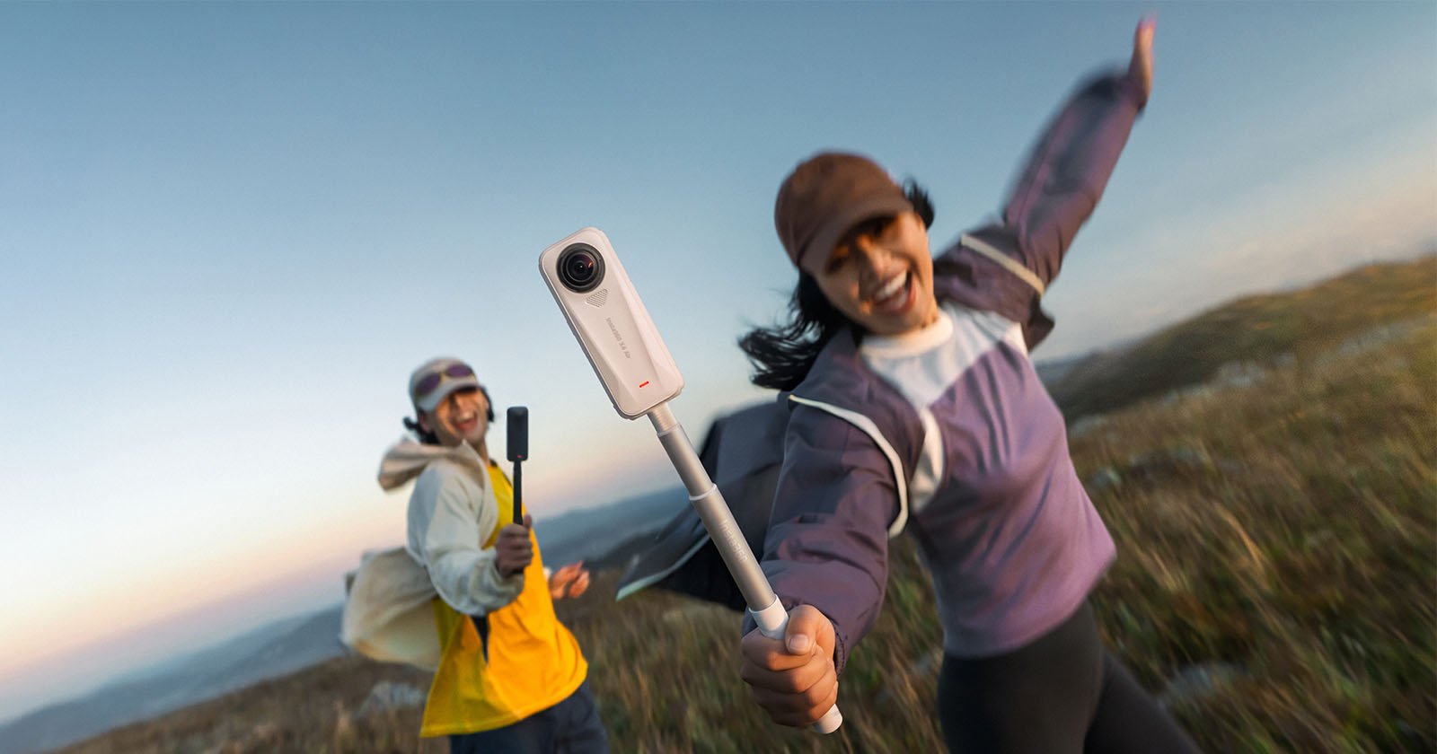Two people are outdoors on a grassy hill. The person in front, smiling and wearing a purple jacket and cap, holds a 360-degree camera on a stick, while the other person, also smiling, walks behind holding another camera.