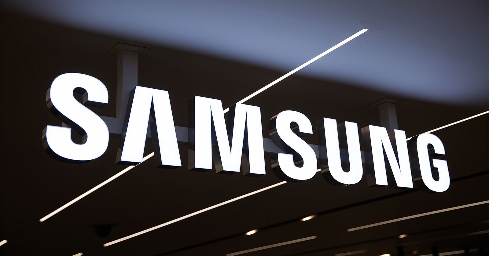 Large illuminated SAMSUNG logo sign on a ceiling, with modern linear lights and a minimalist black and white color scheme in the background.