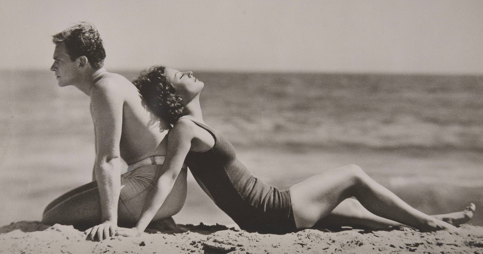 A man and woman in vintage swimsuits sit back to back on a sandy beach, facing the ocean. The woman leans back with her face tilted upward, eyes closed, enjoying the sun. The photo is in black and white.