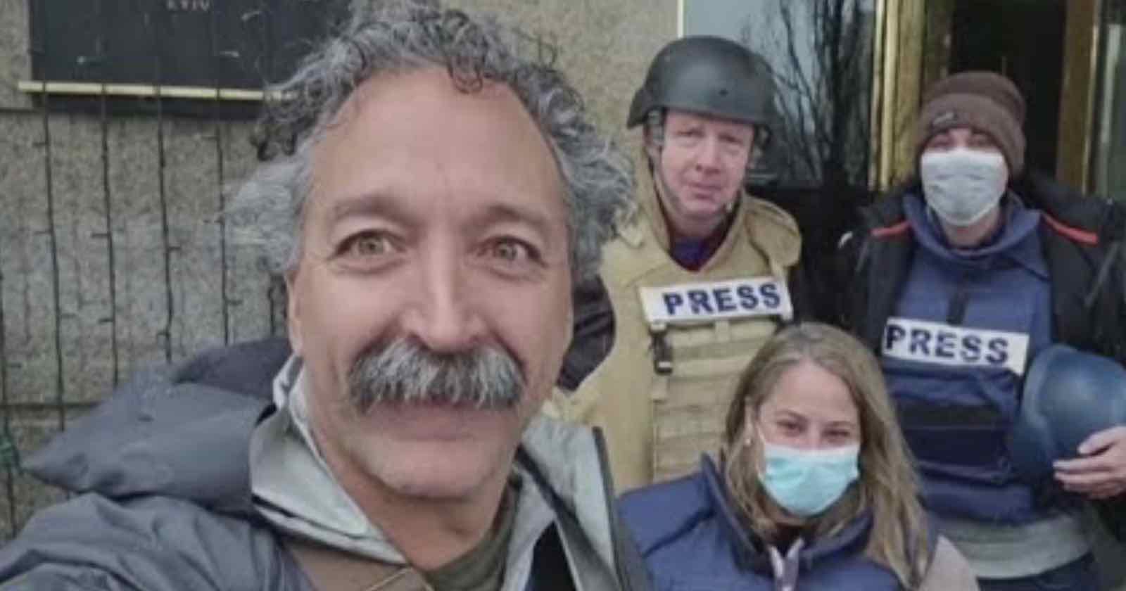 A close-up of four people, three wearing PRESS vests and helmets. Two are standing and one is kneeling. The person in front has curly hair and a mustache, smiling at the camera. Two others are wearing masks, and one holds a helmet.