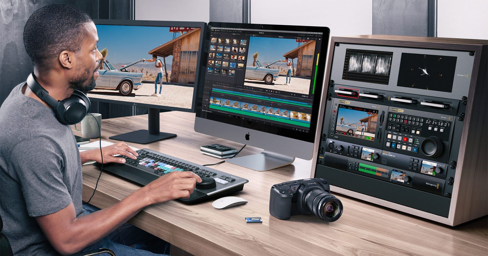 A man sits at a desk editing a video on a large computer; he wears headphones and uses a keyboard with colored keys. Nearby are a camera, an SD card, control panels, and two monitors displaying video footage.
