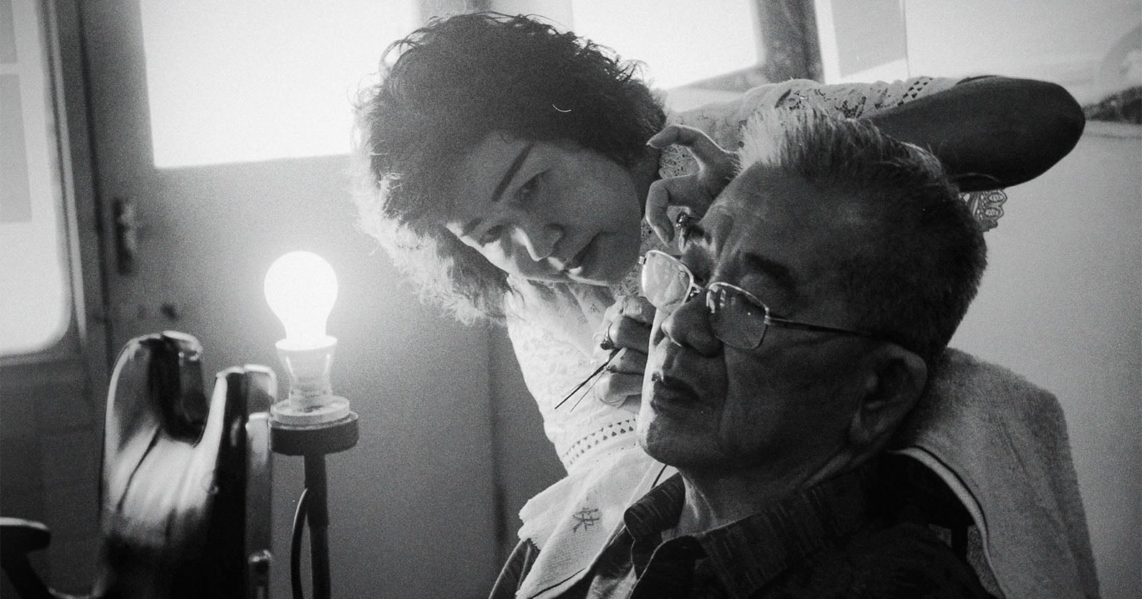 A woman carefully cleans an elderly man's ear as he sits in a chair near a lit lamp in a softly lit room. The scene is captured in black and white.