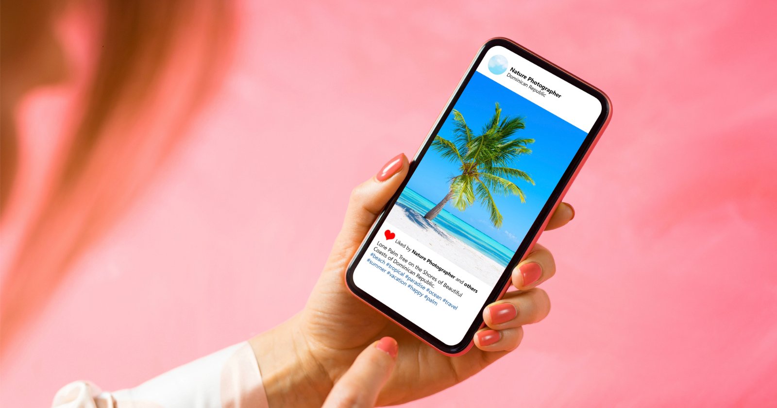 A person with neatly manicured nails holds a smartphone, viewing a social media post featuring a palm tree on a tropical beach under a clear blue sky, against a pink background.