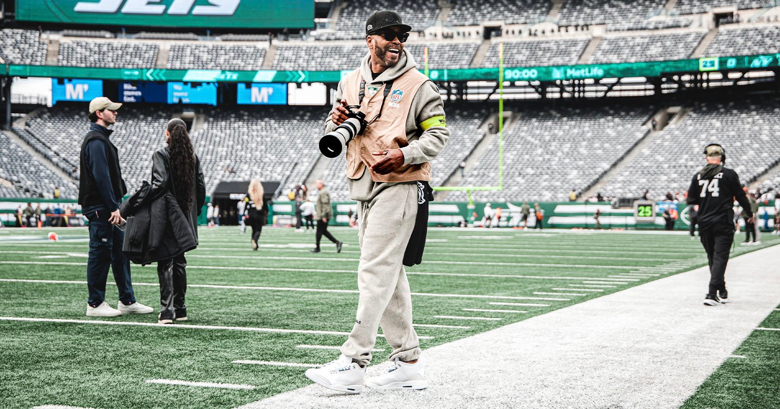 A smiling photographer in casual clothes and a cap stands on the sidelines of a football field holding a camera, with empty stadium seats and a few people in the background.