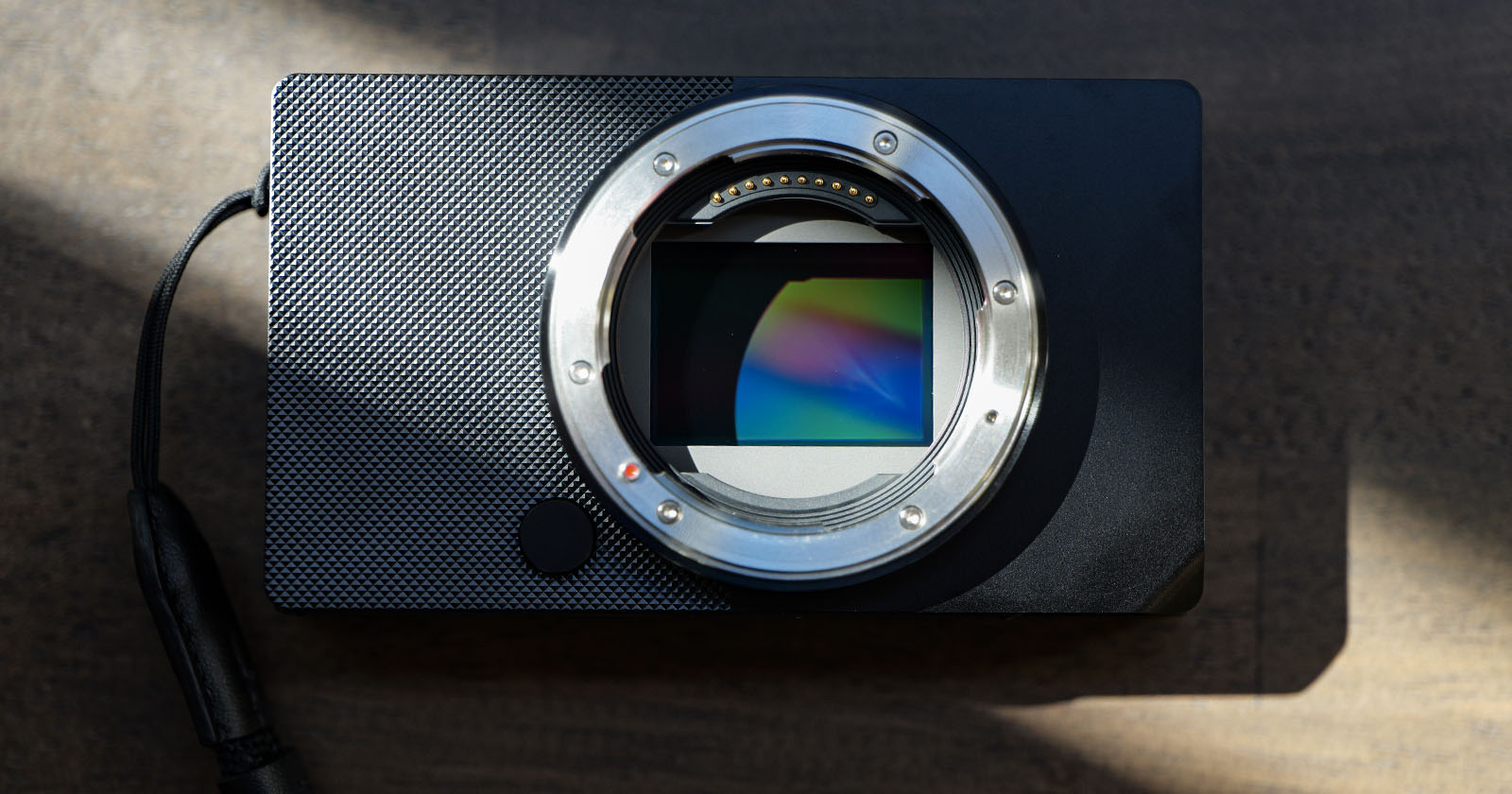 A digital camera body without a lens, showcasing its exposed sensor. The camera is placed on a dark surface with light and shadow patterns across its body, creating a dynamic visual effect. A wrist strap is attached to the left side.