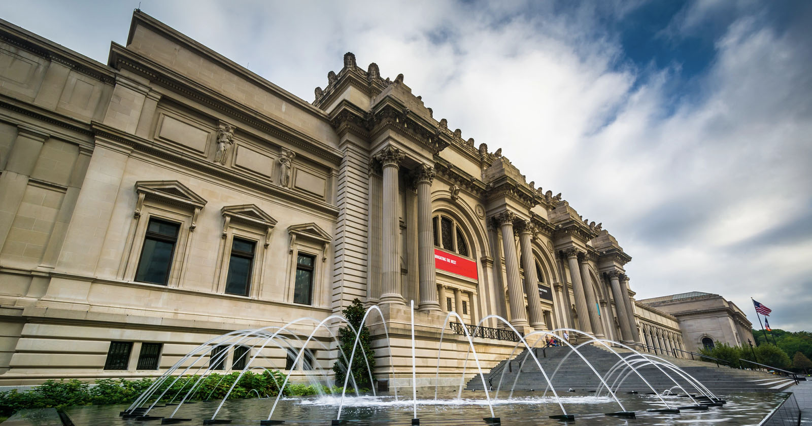 A grand neoclassical building with columns, stone statues, and a wide staircase leading to entrances; water fountains are in front, and a U.S. flag is visible to the right under a partly cloudy sky.