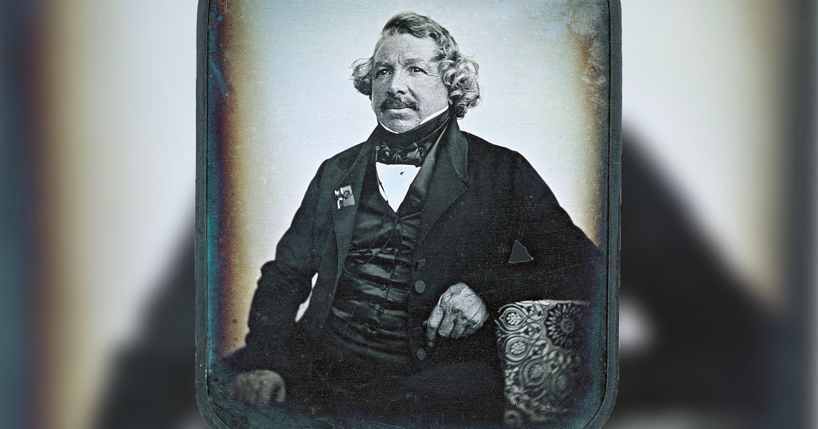 A historical black-and-white portrait of an older man with curly hair, wearing a formal suit and bow tie. He is seated, resting one hand on a patterned cushion. The image background is softly blurred.