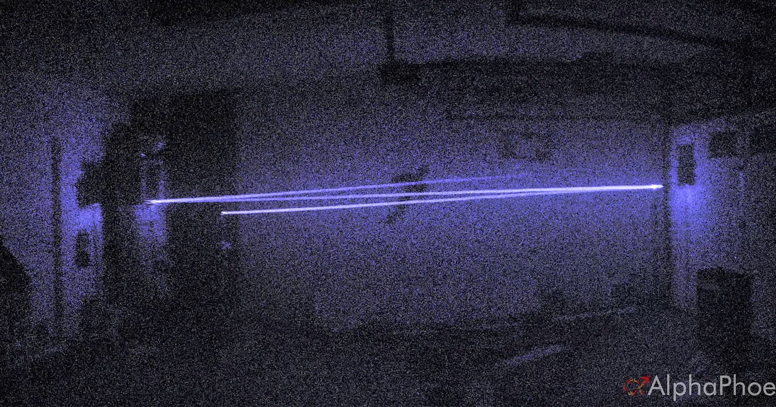 A dark, grainy room with two parallel blue laser beams shining horizontally across the scene. The light creates a glowing effect in the low-light environment.