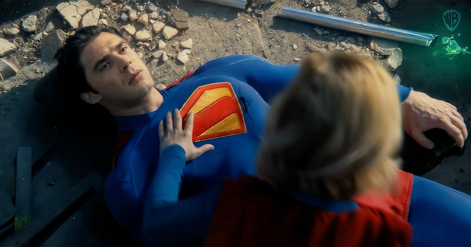 A person in a superhero costume with a large S emblem lies on the ground, surrounded by debris. Another person in a red cape is kneeling beside them, placing a hand on their chest. The scene appears dramatic and intense.