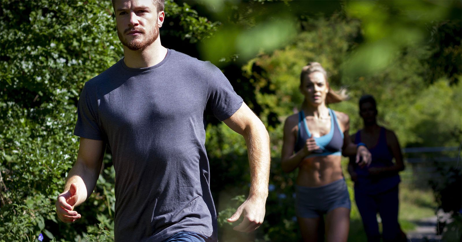 A man in a gray T-shirt jogging on a path lined with trees, followed by two women. The woman directly behind wears a blue sports bra and shorts, while the other is in darker workout clothes. Sunlight filters through the leaves, casting soft shadows.