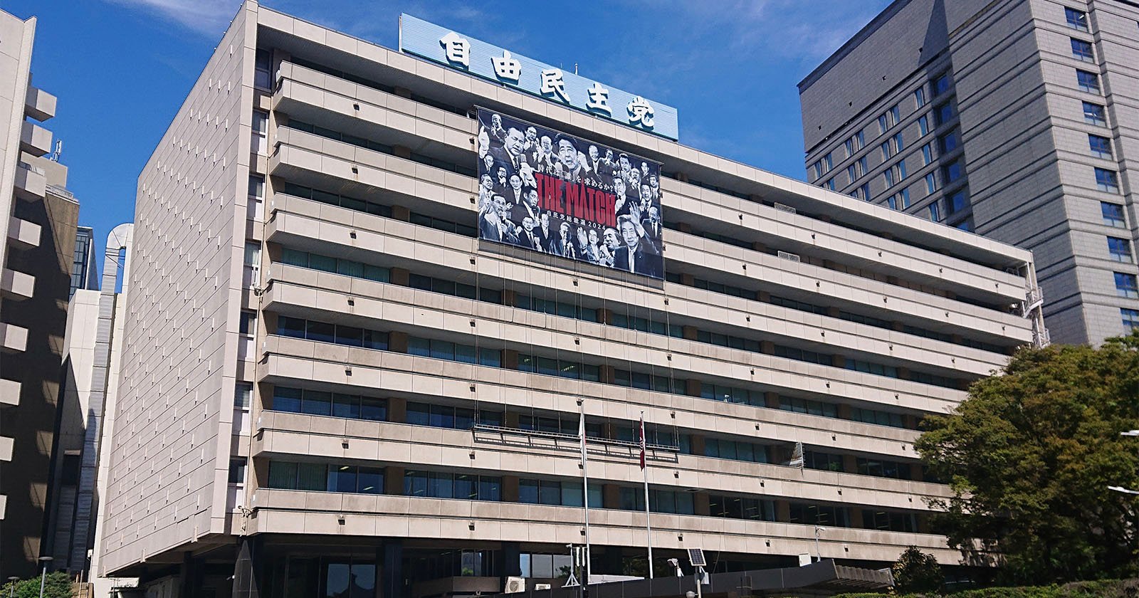 A large modern office building with a Japanese sign on the roof and a black-and-white banner featuring characters from the anime Attack on Titan displayed across its facade. Blue sky and other tall buildings are in the background.