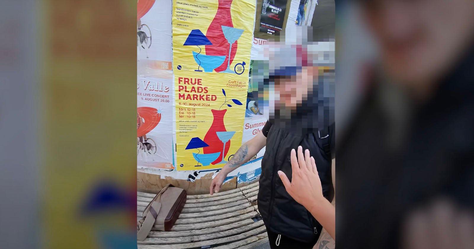 A person in a black vest and cap stands in front of colorful posters, one of which reads FRUE PLADS MARKED. A hand, partially visible in the foreground, seems to reach towards the person. There is a suitcase or bag on a bench nearby.