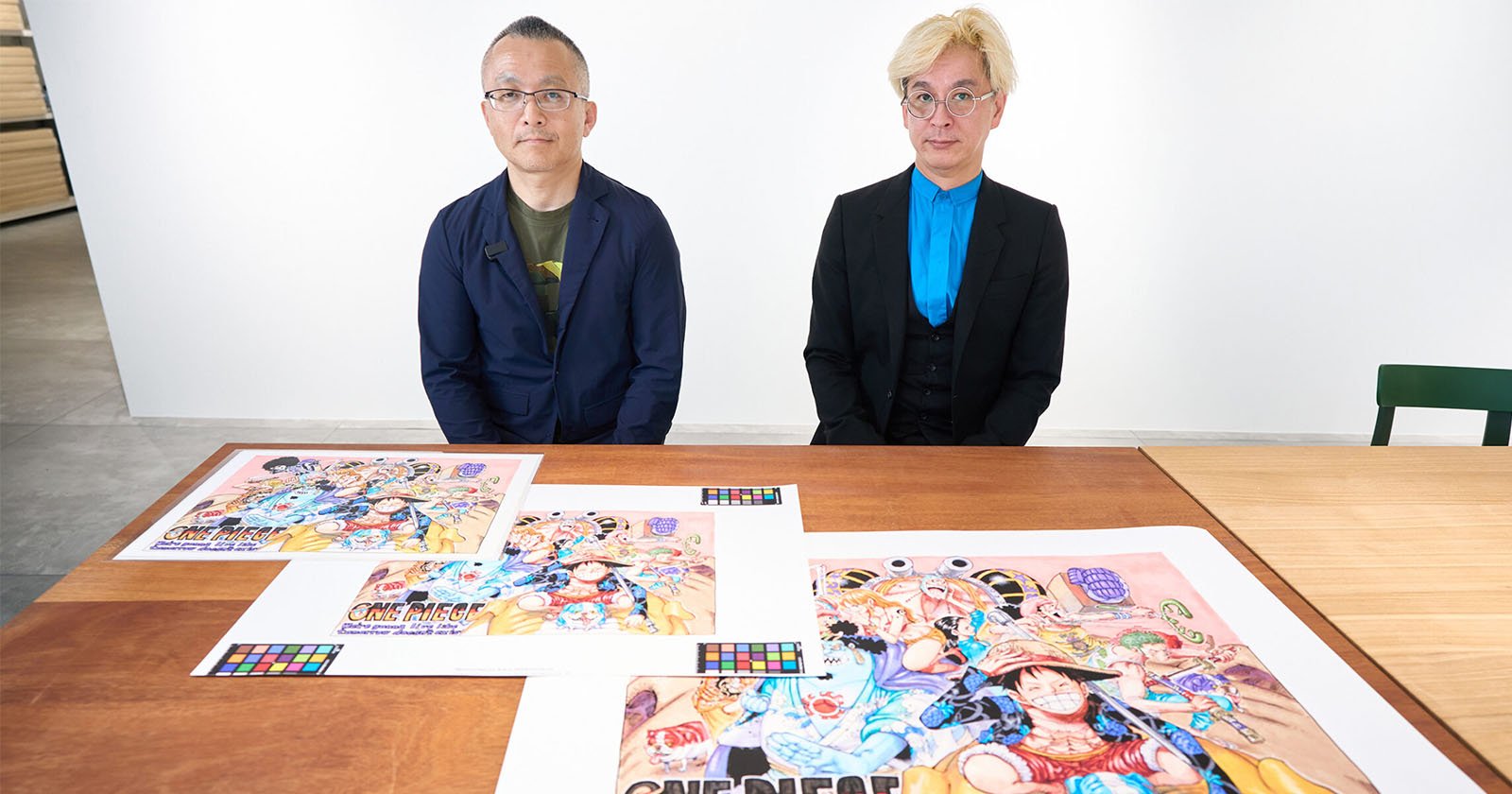 Two men sit at a table with colorful One Piece anime artwork spread out in front of them, against a plain white background. Both men are wearing glasses and formal jackets.